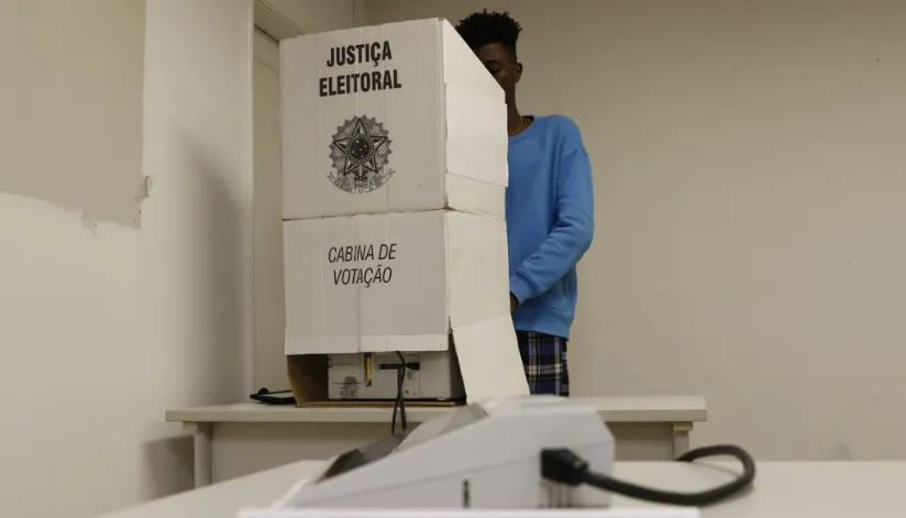 A foto mostra uma cabine de votação. Uma pessoa vestida de azul está atrás dela, votando.