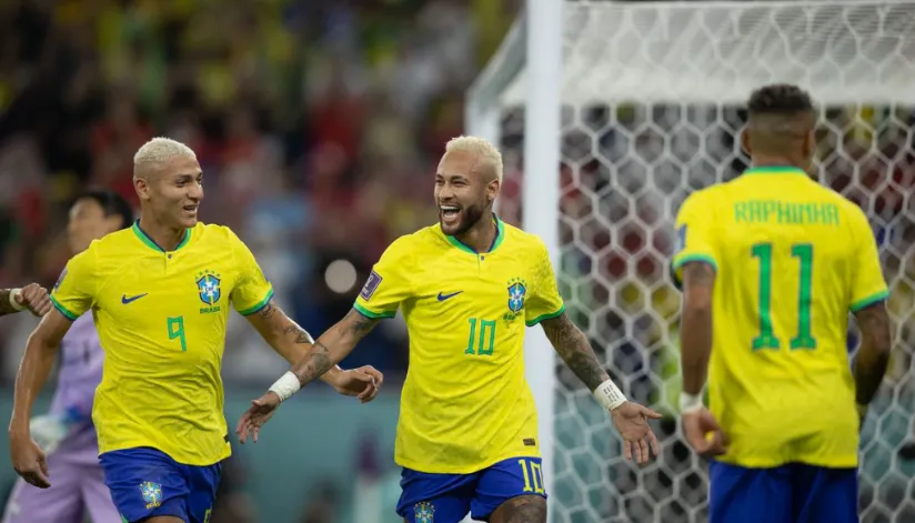 A foto mostra os jogadores Neymar e Richarlison comemorando. O jogador Raphinha está levemente desfocado e de costas para a câmera.